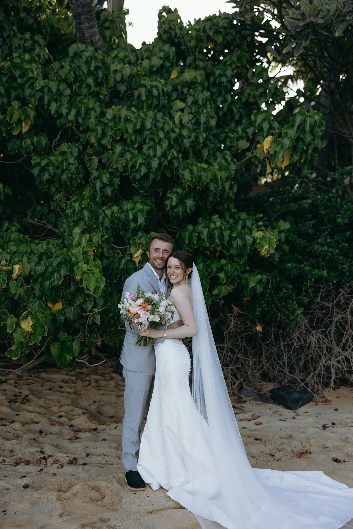 Newlyweds smiling together in front of tropical greenery in romantic sneak peek photos