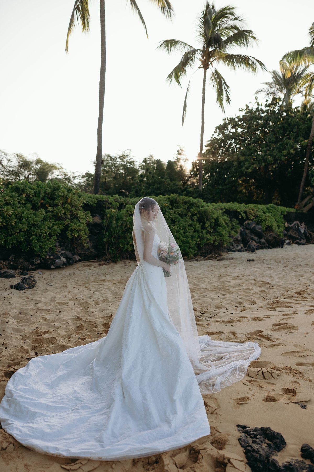 Bride standing alone with long veil flowing on beach during golden hour in wedding sneak peek photos