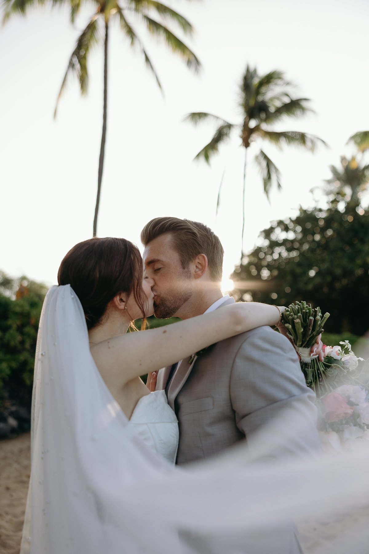 Golden hour kiss between bride and groom under palm trees in romantic sneak peek photos