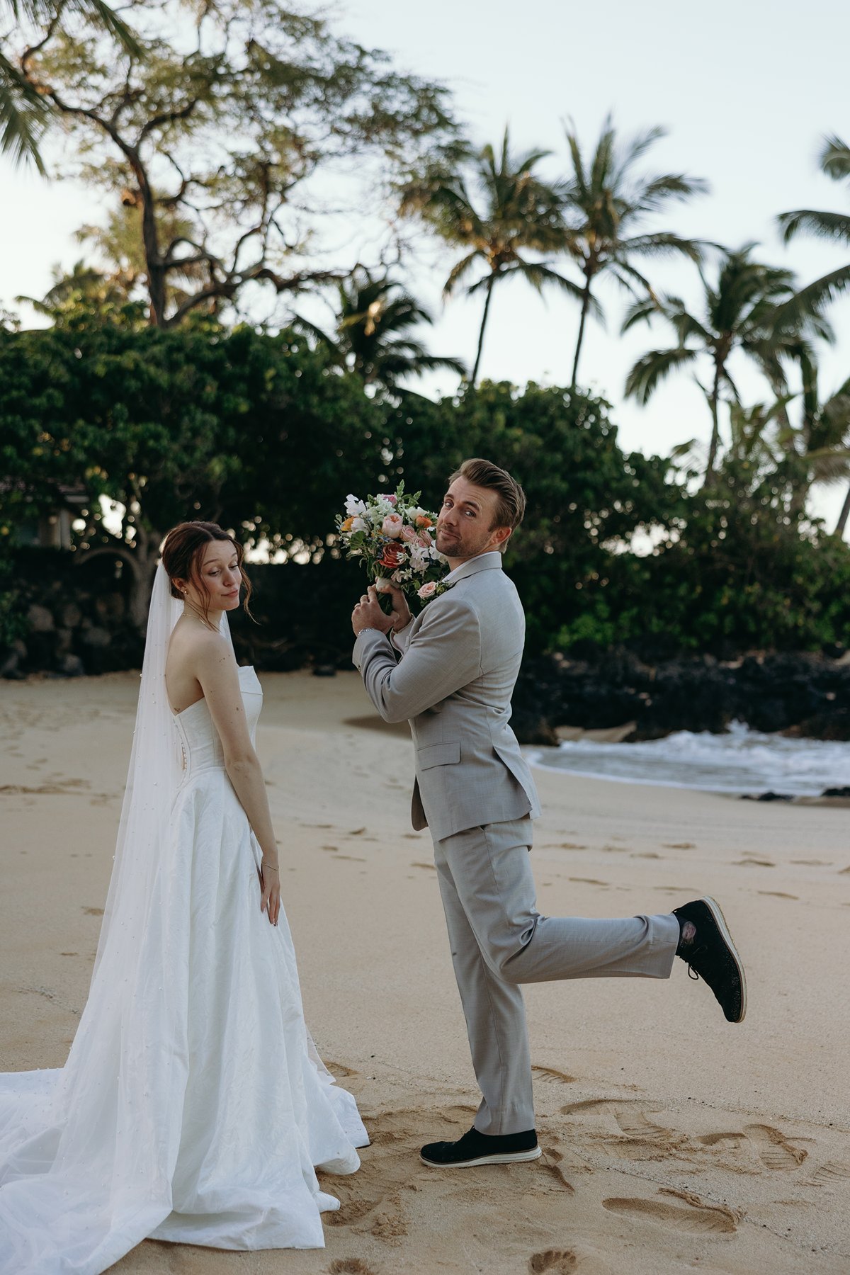 Playful groom posing with bouquet beside bride on the beach in fun wedding sneak peek photos