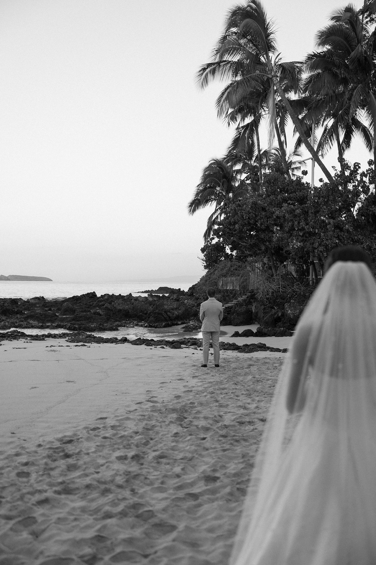 Artistic black and white sneak peek photos of bride approaching groom for first look on beach
