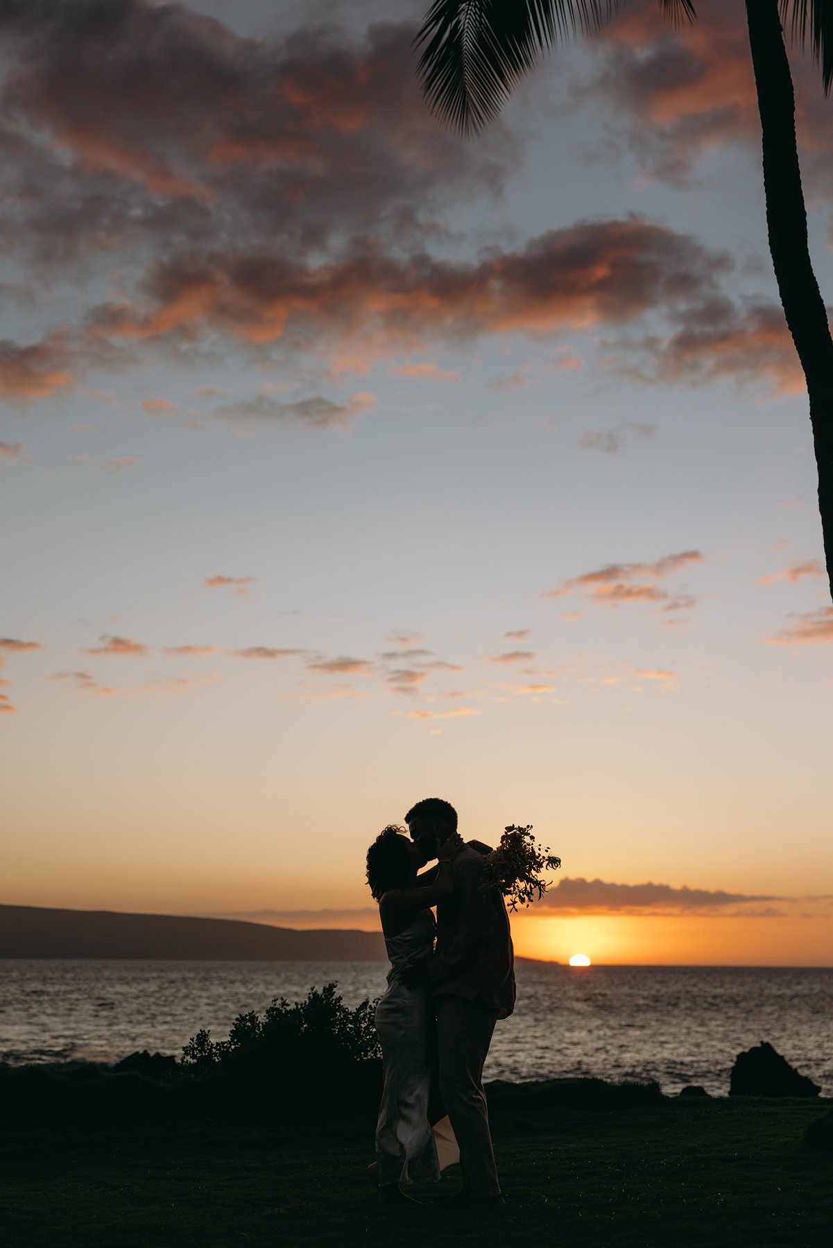 Newlyweds sharing a kiss as the sun sets over the ocean during an intimate Hawaii wedding celebration