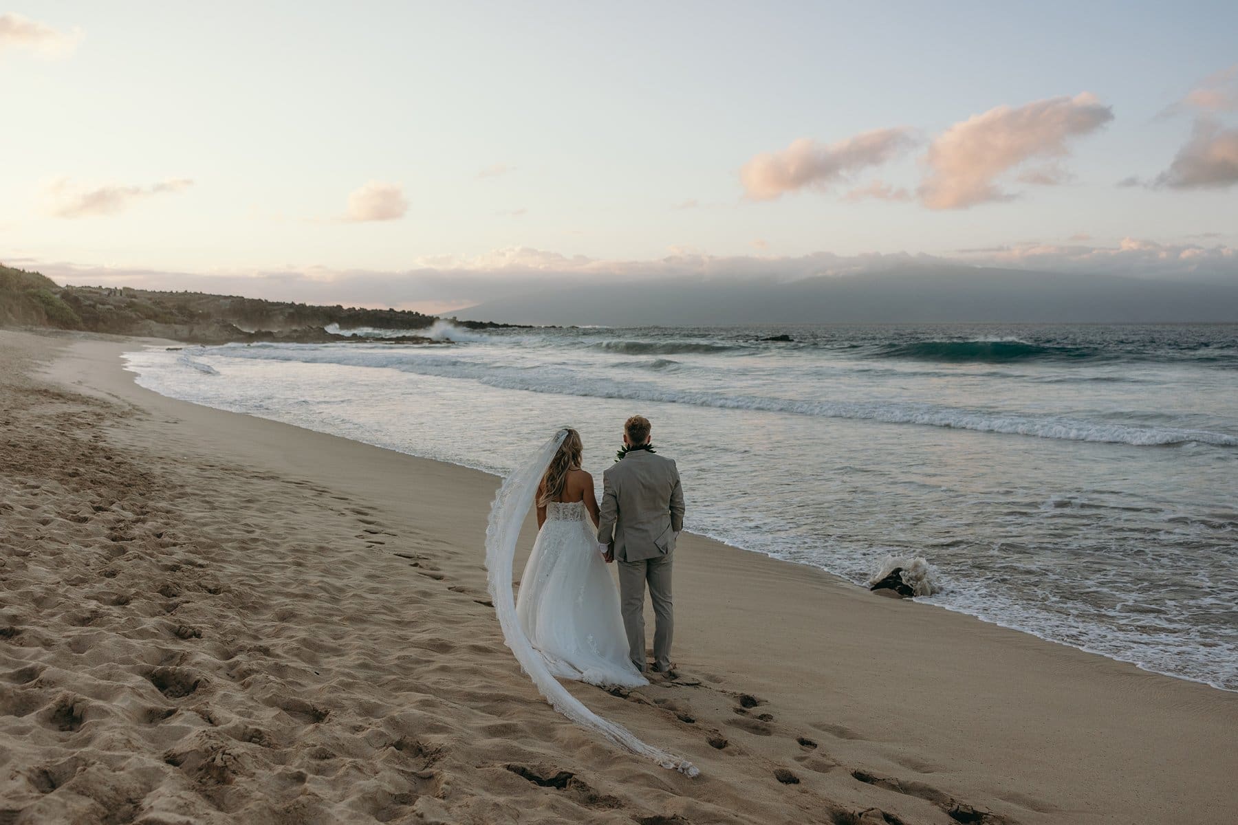 Bride and groom walking hand in hand along a quiet beach at sunset during a Maui elopement
