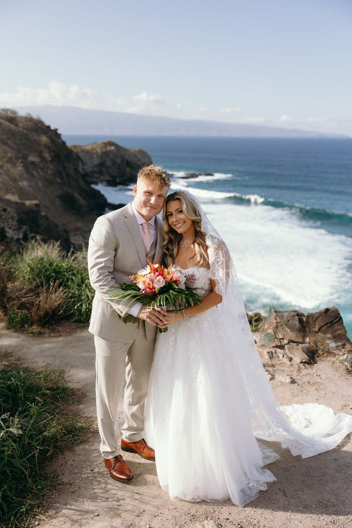 Newlyweds posing above dramatic coastal cliffs with ocean views stretching across Maui