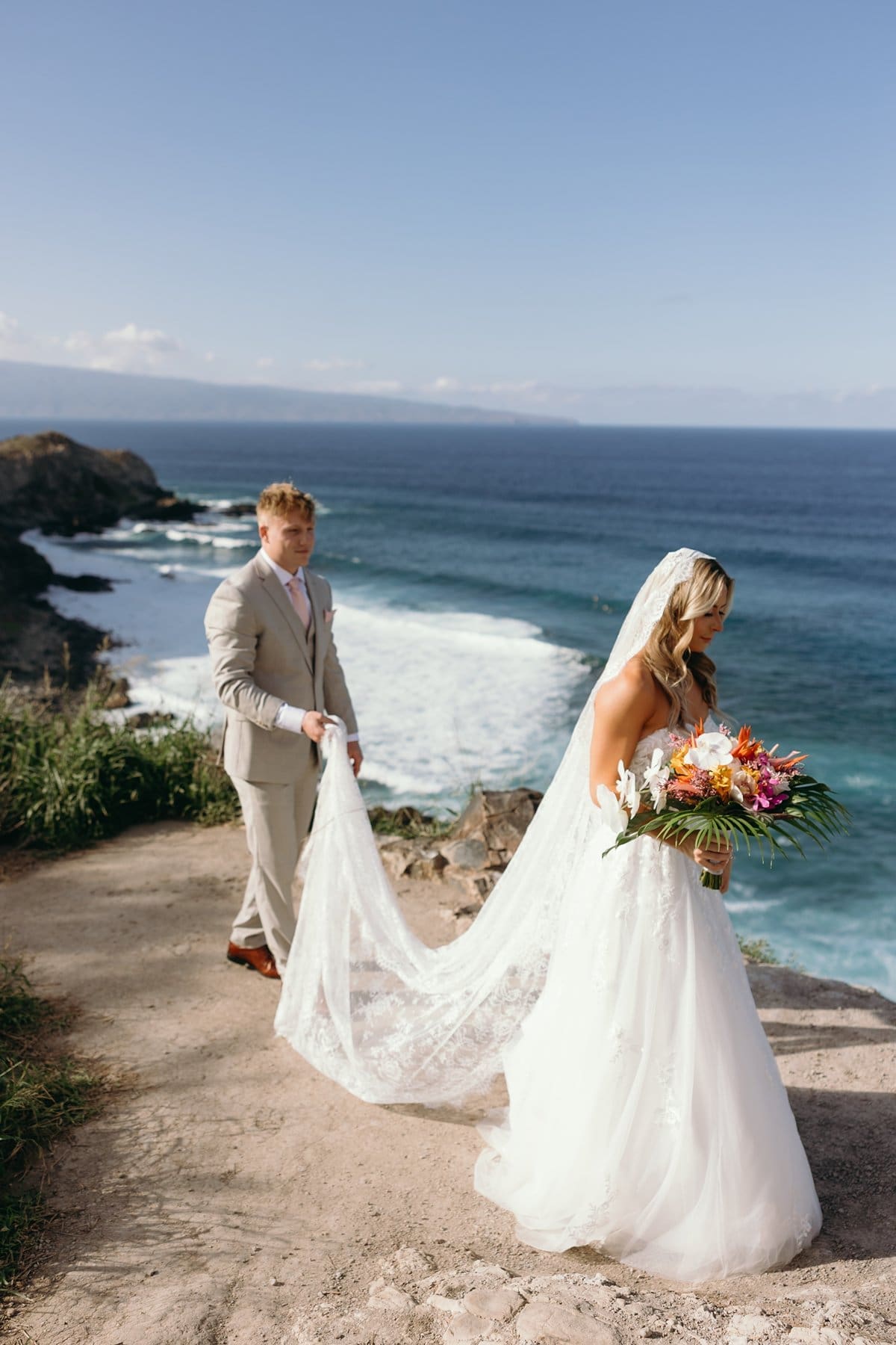 Groom carrying the bride's veil along a coastal overlook during sunset portraits after their Maui elopement